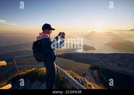 Junger Mann fotografiert Landschaft mit See und Bergen bei schönem Sonnenaufgang. Blick vom Pilatus-Berg, Luzern, Schweiz Stockfoto