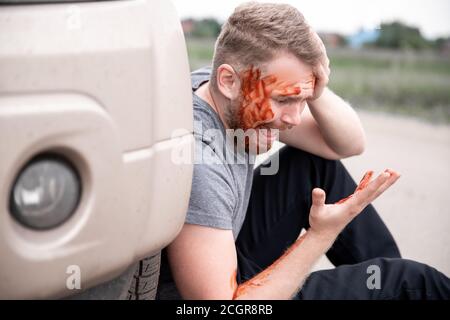 Der junge Mann sitzt mit blutigem Kopf in der Nähe des Wagens, schreiend und bereutend handeln Stockfoto