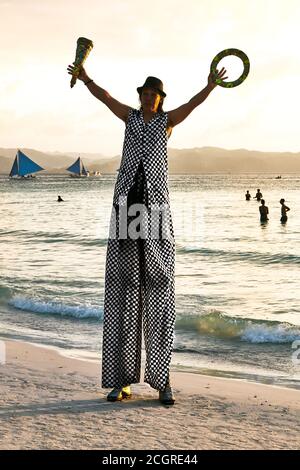 Mann auf Stelzen beim Jonglieren am Weißen Strand bei Sonnenuntergang, mit lokalen Touristen als Zuschauer, nur wenige Menschen schwimmen und blaue Segelboote als Hintergrund Stockfoto