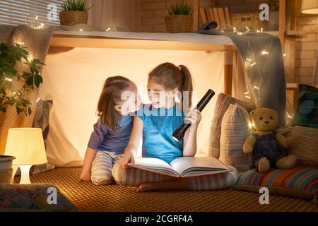 Zwei niedliche kleine Kinder lesen ein Buch mit Taschenlampen im Zelt. Glückliche Mädchen spielen zu Hause. Lustige, nette Kinder, die Spaß im Zimmer haben. Stockfoto