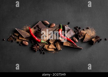 Dunkler Schokoladenhintergrund. Schokoladenstücke, Kakaopulver, scharfe Chilischoten und Gewürze auf schwarzem Tisch, Draufsicht, Kopierraum. Stockfoto
