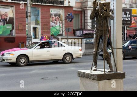 Denkmal für den Fotografen vor dem Hintergrund von Hochzeitsautos auf einer Stadtstraße. Sehenswürdigkeit befindet sich im Zentrum von Krasnojarsk, Krasnojarsk Terri Stockfoto