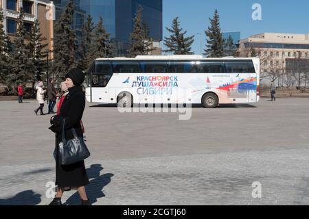 Eine ältere Frau wartet auf einen zweistöckigen Tourbus mit der Aufschrift "Krasnojarsk - die Seele und Macht Sibiriens" auf dem Stadtplatz. Stockfoto