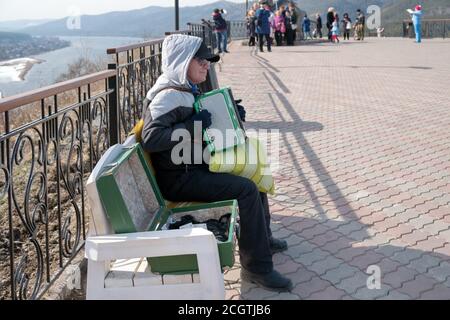 Ein Mann verdient Geld, spielt Akkordeon und sitzt auf einer Bank auf der Aussichtsplattform über dem Fluss Jenisei. Stockfoto