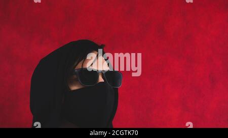 Mann in Schutzmaske und Sonnenbrille. Schwarze Kleidung mit Kapuze. incognito auf rotem Hintergrund. Kopierraum Stockfoto