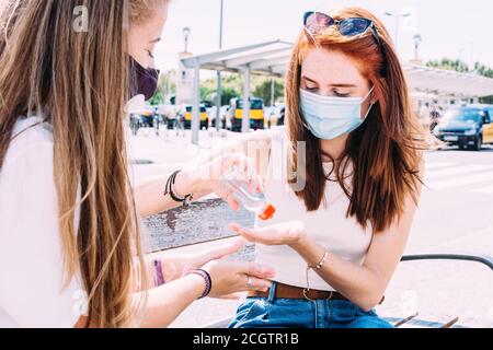 Zwei junge Frauen sitzen auf einer Bank vor dem Zug Station auf antibakterielles Gel setzen Stockfoto