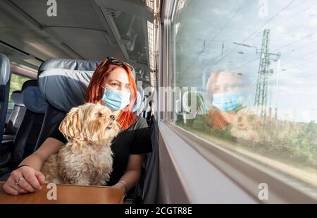 Junge Rotschopf Frau und Hund mit dem Zug unterwegs, während Pandemie. Millennial Mädchen mit einer medizinischen Maske auf einem deutschen Fernverkehrszug, in der Business Class. Stockfoto