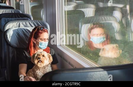 Junge rothaarige Frau mit Gesichtsmaske, die mit dem Zug unterwegs ist, in erster Klasse. Mädchen und Hund reisen mit dem deutschen Intercity-Expresszug. Pandemie-Sicherheit auf Reisen. Stockfoto