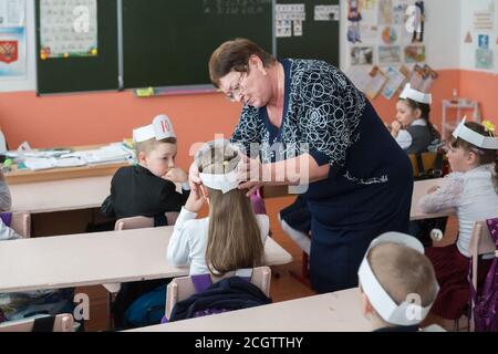 Die Lehrerin stellt den Hut auf das Schulmädchen, das an ihrem Schreibtisch sitzt, vor den Hintergrund des Schulrates in der Klasse der ländlichen Grundschule. Stockfoto