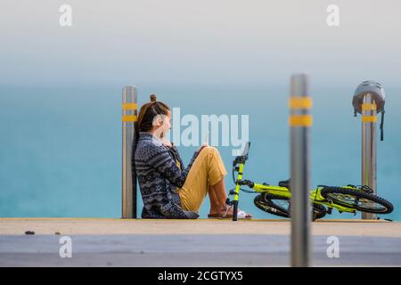 Moment des Restes. Mutter mit Handy sitzt am Rande des Meeres in der Nähe ihres Kinderfahrrads. Stockfoto