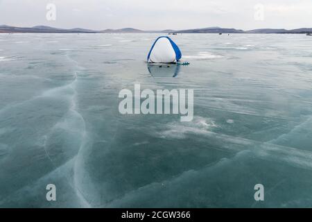 Ein Fischerzelt steht auf dem Eis eines Schmelzsees, bedeckt mit Rissen, im frühen Frühjahr. Stockfoto