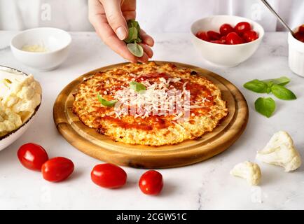 Frauenhände bestreuen frische Basilikumblätter auf der Oberseite der Blumenkohlpizzenkruste. Keto Diät koncept. Gesunde Ernährung Stockfoto