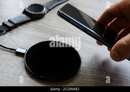 Das in der Hand gehaltene Smartphone wird auf das kabellose Ladegerät gestellt. Intelligente Uhr im Hintergrund Stockfoto