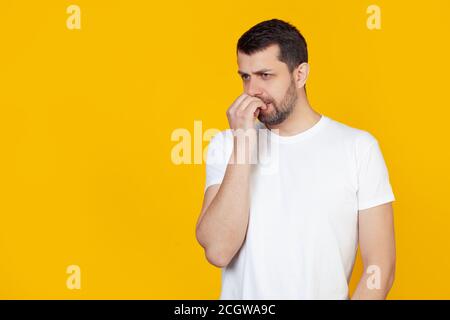 Ein moderner junger Mann mit einem Bart in einem weißen T-Shirt sieht angespannt und nervös aus, wenn seine Hände auf dem Mund seine Nägel beißen. Das Problem der Angst, auf einem gelben Hintergrund Stockfoto