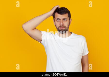 Ein junger Mann mit Bart in einem weißen T-Shirt, besorgt über ein Problem, mit der Hand auf dem Kopf, nervös und besorgt über die Krise, steht auf einem isolierten gelben Hintergrund. Stockfoto