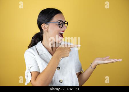 Schöne Frau über gelben Hintergrund überrascht, zeigt und zeigt etwas, das auf ihrer Hand ist Stockfoto