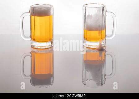 Bierbecher mit Bier. Bier ohne Zuckerguss. Bierbecher auf weißem Hintergrund mit Hintergrundbeleuchtung. Ein Glas fast leer und eines voll. Stockfoto