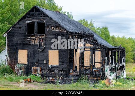 Ein verbranntes Haus. Die Außenseite des Gebäudes ist komplett schwarz verkohlt, mit Ausnahme von Sperrholz, das einige Fenster bedeckt. Bäume dahinter und wolkigen Himmel. Stockfoto