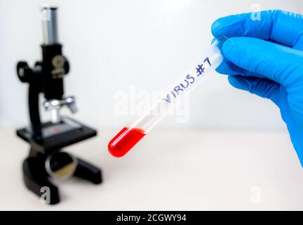 Eine Hand trägt einen blauen Handschuh und hält ein Reagenzglas mit VIRUS #7 im Marker. Das Reagenzglas enthält eine kleine Menge roter Flüssigkeit. Mikroskop im Bac Stockfoto