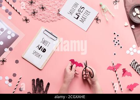 Halloween-Textkarten mit Text Sweet and Spooky und Skelett lebt Materie. Hände mit Schere schneiden Papierdekorationen auf rosa Hintergrund. Konfetti, Pap Stockfoto
