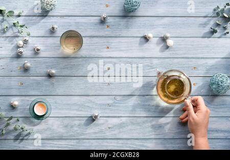 Wintertee, flache Lay-Anordnung mit Glas Teekanne, Glas Tee in der Hand auf verblassten blauen Holzplanken. Weihnachtsschmuck auf dem Tisch - Spiegel Stockfoto