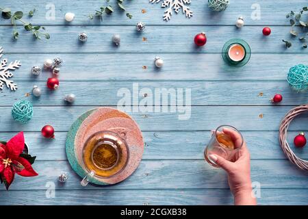 Wintertee, flache Lay-Anordnung mit Glas Teekanne, Glas Tee in der Hand auf verblassten blauen Holzplanken. Weihnachtsdekorationen - Spiegelkugeln, Kugel Stockfoto