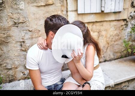 Liebespaar küsst sich hinter weißem Frauenhut auf der Steinbank in der Altstadt. Junger Mann und Frau umarmen. Sommer sonnigen Tag Stockfoto