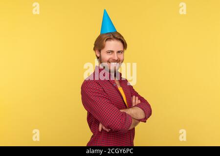 Happy guy mit lustigen Party-Kegel auf Kopf stehen gekreuzte Hände, Blick auf die Kamera mit toothy Lächeln, Geburtstag feiern, Jubiläum. Indoor Stockfoto