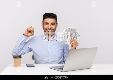 Positive elegante Geschäftsmann sitzen Büroarbeitsplatz mit Laptop, lächelnd hält Geld Dollar und goldenen Bitcoin btc, Kryptowährung Investition. In Stockfoto