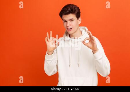 Alles ist in Ordnung. Stilvoller Teenager in weißem Kapuzenpullover, der eine gute Geste und ein Augenzwinkern zeigt. Genehmigung des Antrags der Universität. Innenaufnahmen im Studio isoliert auf oder Stockfoto