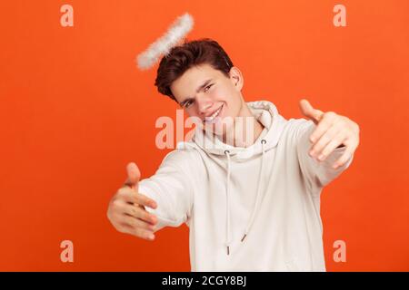 Kommen Sie hierher. Schöner freundlicher Mann im lässigen Hoodie mit Nimbus über dem Kopf, der vorgibt, Engel zu sein und die Hände bereit hält, sich zu umarmen. Innenaufnahme des Studios isola Stockfoto