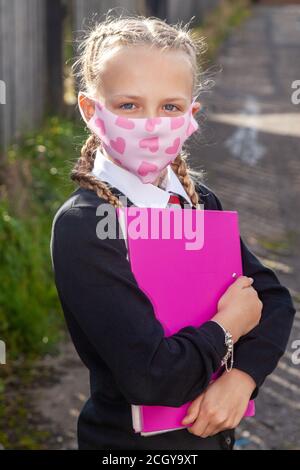 Eine zehnjährige Schülerin mit ihren Haaren in Zöpfen, die einen pinken A4-Ordner in der Hand halten und eine Gesichtsmaske tragen. Stockfoto