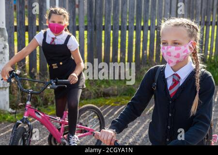 Zwei zehnjährige Schulfreunde in Schuluniformen sitzen auf Fahrrädern und tragen Gesichtsmasken. Stockfoto