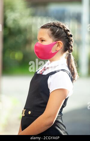 Eine zehnjährige Schülerin in Schuluniform mit ihren Haaren in Zöpfen und einer rosa Gesichtsmaske. Stockfoto