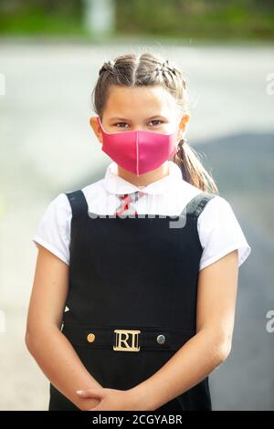 Eine zehnjährige Schülerin in Schuluniform mit ihren Haaren in Zöpfen und einer rosa Gesichtsmaske. Stockfoto