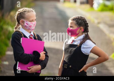 Zwei 10-jährige Schülerinnen in Schuluniform und Gesicht Masken stehen draußen und schauen sich an Stockfoto