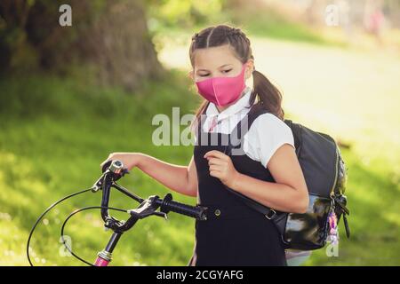 Eine zehnjährige Schülerin, die mit der Hand auf dem Fahrrad steht und eine Gesichtsmaske trägt. Stockfoto