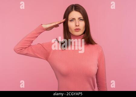 Ernst fleißig Brünette Frau in rosa Pullover Hand in der Nähe des Kopfes zeigt Gruß Geste, Disziplin. Innenaufnahme des Studios isoliert auf pinkfarbenem Hinterteil Stockfoto