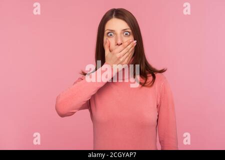 Schockiert verängstigte Frau mit braunen Haaren Mund schließen mit Hand Blick auf Kamera mit großen Augen, halten Stille. Innenaufnahme des Studios isoliert auf rosa Bac Stockfoto