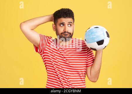 Trauriger nachdenklicher Mann im roten T-Shirt, der den Fußball in Schutzmaske hält, Symbol des Fußballwettbewerbs während des covid-19 Virus, Score-Fälschung. ICH Stockfoto