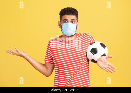 Frustriert unsicheren Mann in roten T-Shirt und hygienische Gesichtsmaske Verbreitung Hände halten Ball, verwirrt, wie Fußball Sicherheit zu spielen. Innenaufnahme im Studio Stockfoto