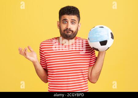 Frustrierter junger bärtiger Mann im roten T-Shirt, der in der Hand Fußball in medizinischer Maske hält, Probleme mit der Fußballmeisterschaft, Korruption. Innen s Stockfoto