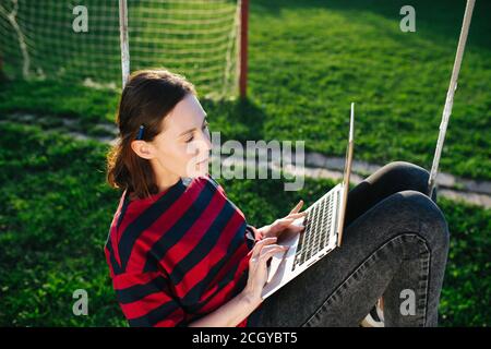 Friedliche junge Frau, die auf den Schaukeln auf einem Rasen sitzt Mit Laptop Stockfoto