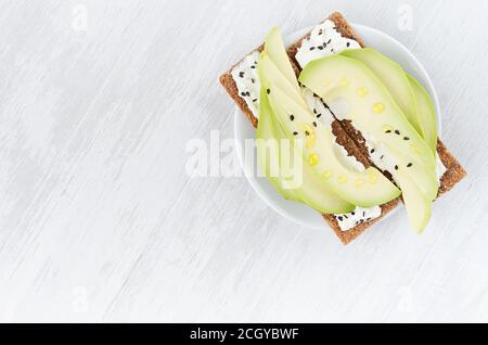 Bio-Season Sandwiches auf trockenem braunem Korn-Knäckebrot mit frischen Avocado-Scheiben, Olivenöl, schwarzen Sesam-Samen, Frischkäse auf weißem Holztisch, Stockfoto