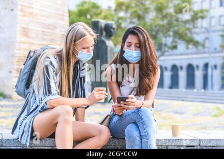 Zwei junge schöne Freundinnen sitzen auf einer Steinbank Auf der Straße und tragen blaue Hygienemasken zeigen jeden Bildschirm für anderes Smartphone Stockfoto