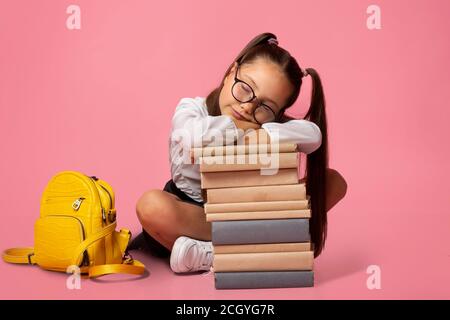 Trauriges und müdes Konzept. Mädchen in Brille in Uniform mit Rucksack schläft auf Stapel von Büchern Stockfoto