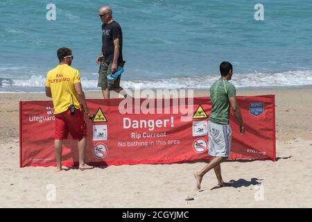 Urlauber, die an einem RNLI-Rettungsschwimmer vorbeilaufen und ein Warnschild am Fistral Beach in Newquay in Cornwall aufhängen. Stockfoto