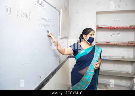 Weibliche Lehrerin Tragen Maske und lehren zeigen auf Whiteboard im Klassenzimmer, indische Schulklasse während covid19 Pandemie, neue normale, Kopie Spa Stockfoto