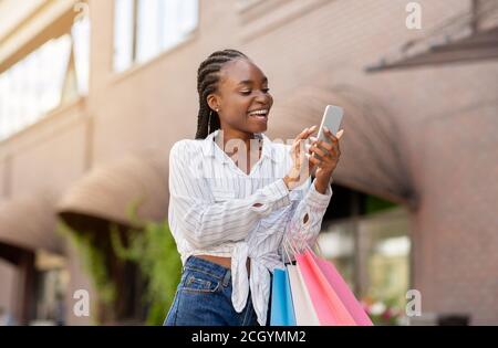 Sonderangebot für Stammkunden. Lachende afroamerikanische Mädchen mit Taschen erhielt Nachricht am Telefon Stockfoto