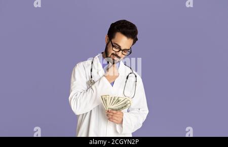 Korruption in der medizinischen Industrie. Indischer Arzt hält Fan von Geld auf lila Hintergrund Stockfoto
